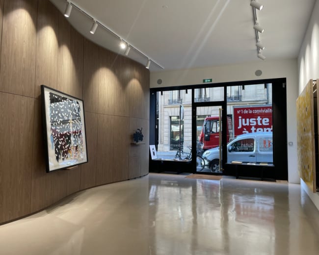 The image shows the interior of a modern art gallery with wooden paneling, featuring several artworks on the walls and large windows facing a street.