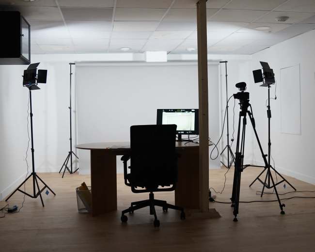 A minimalist office space features a desk with a computer, an office chair, and two lighting setups on tripods, accompanied by a backdrop for recording.
