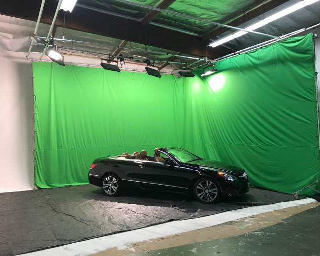 A black convertible car is positioned on a black tarp in front of a green screen setup inside a studio.