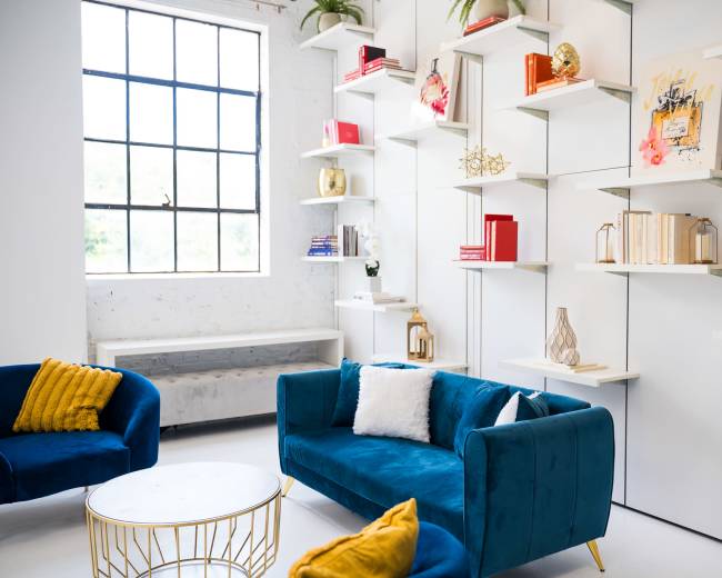 The image shows a modern interior living space featuring a bright window, a stylish blue sofa, two blue chairs, and a decorative shelving unit displaying books and decorative items.