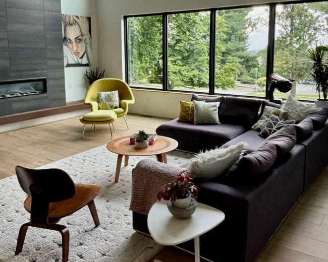 A modern living room with large windows overlooking greenery, a sectional sofa, a round coffee table, and a mix of decorative plants and art.