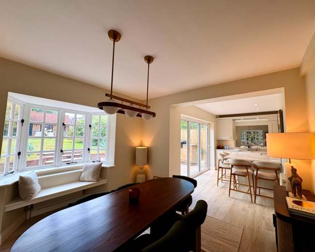 A dining area features a long table with a chandelier overhead, and sunlight streams in through large windows, leading into a modern kitchen visible in the background.