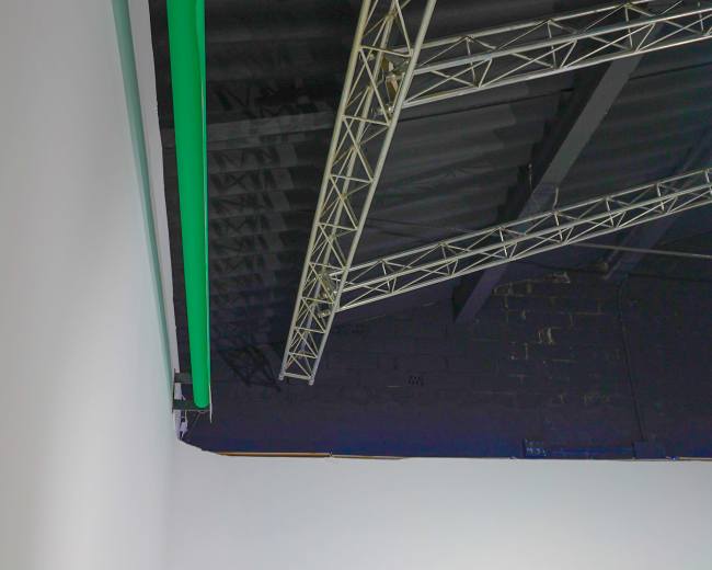 The image shows the corner of a room with a white wall and a ceiling featuring a green light and exposed truss support beams.