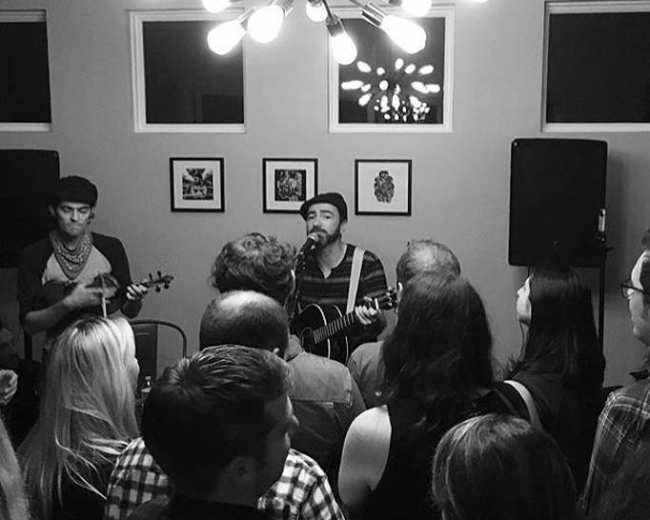 A musician plays guitar and sings to an audience while accompanied by another musician on a small stage in a dimly lit room.
