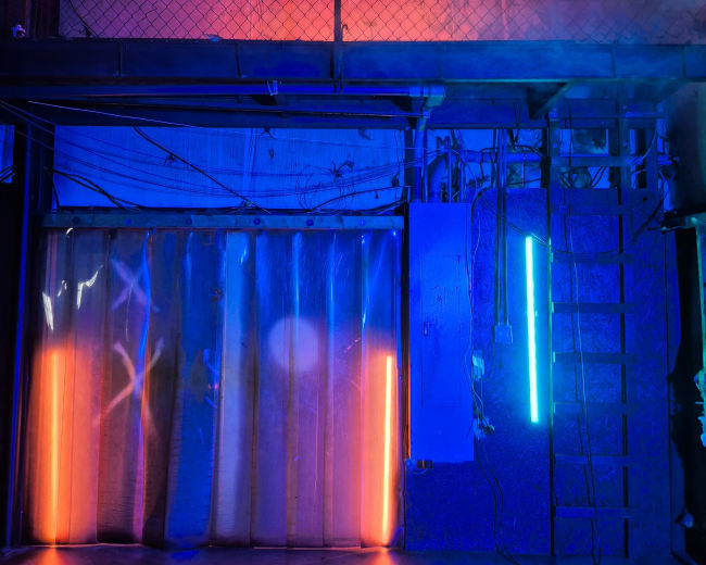 The image shows a dimly lit room with colored lights illuminating a plastic curtain and a metal ladder against a wall.