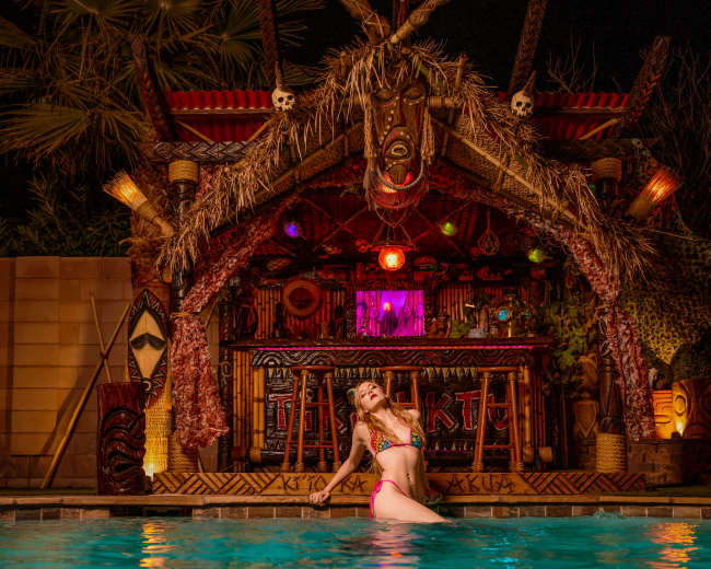 A woman poses by a swimming pool in front of a tiki-style bar decorated with tropical motifs and vibrant lights.