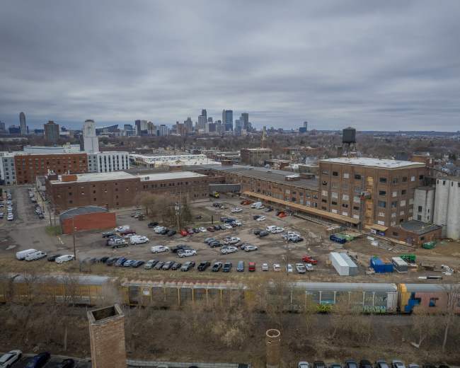 The image shows an aerial view of a large, abandoned industrial area with several buildings and parked vehicles, set against a skyline of a city in the background.