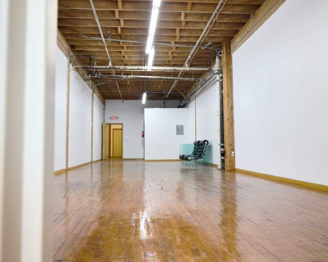 The image shows a spacious, empty room with wooden flooring, high ceilings, and exposed beams, featuring a green chair against one wall and a doorway leading to another area.