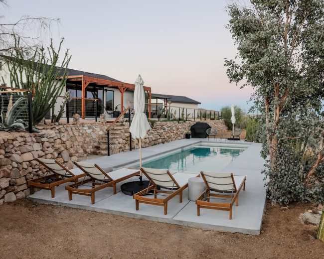 A swimming pool surrounded by wooden lounge chairs and shaded by a large umbrella, set in a desert landscape with stone walls and surrounding vegetation.
