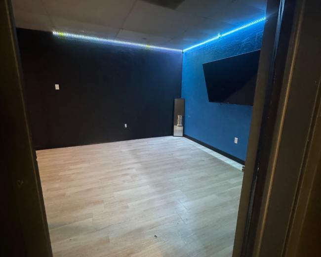 The image shows an empty room with light-colored wooden flooring, black and blue walls, and a mounted television on one wall.
