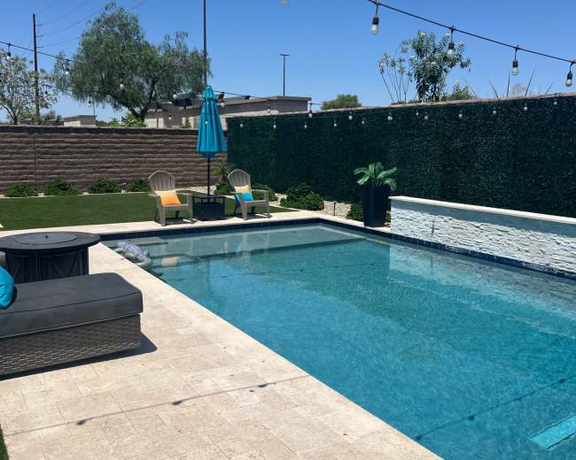 The image shows a clear swimming pool surrounded by a patio with two lounge chairs, an umbrella, and decorative plants against a green wall.