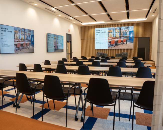 The image shows a modern conference room with long tables arranged in rows, black chairs, and two large screens displaying welcome messages on the wall.