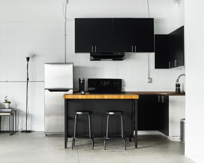 The image shows a modern kitchen with a stainless steel refrigerator, black cabinetry, a wooden countertop with two stools, and a black sofa with a red pillow.