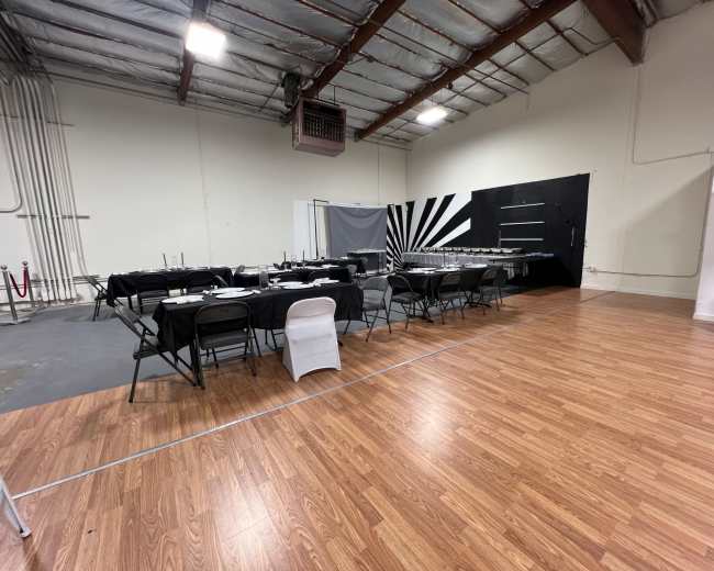 The image shows a spacious indoor area with a set of tables and chairs arranged for dining, featuring a black-and-white themed backdrop and polished wooden flooring.