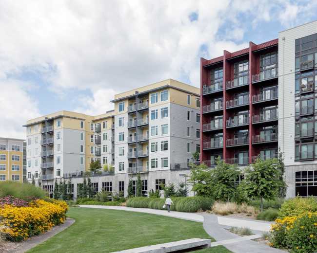 A row of modern apartment buildings with balconies and landscaped gardens are set alongside a paved walking path.