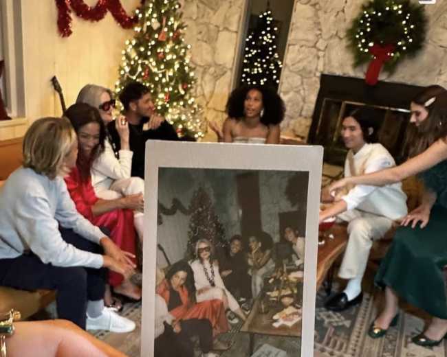 A group of people gathers around a festive setting, with a decorated Christmas tree and a fireplace in the background, while one person holds a polaroid photo of the gathering.