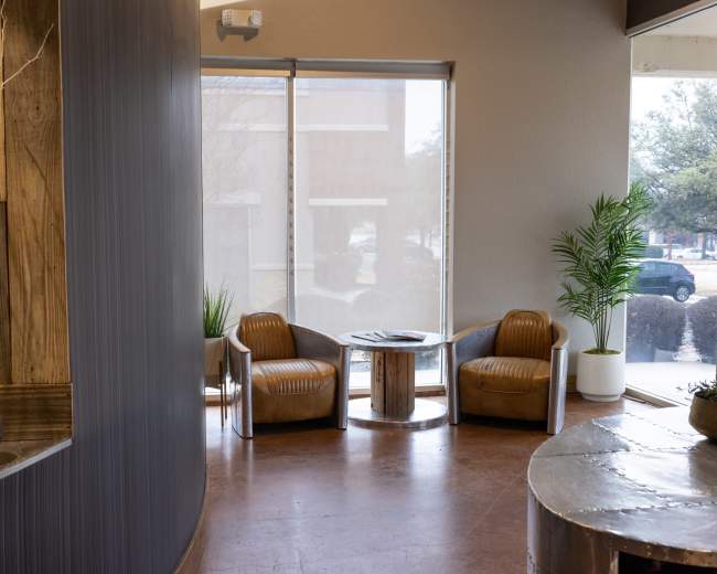 The image shows a modern waiting area featuring two brown chairs, a small table, and large windows letting in natural light.
