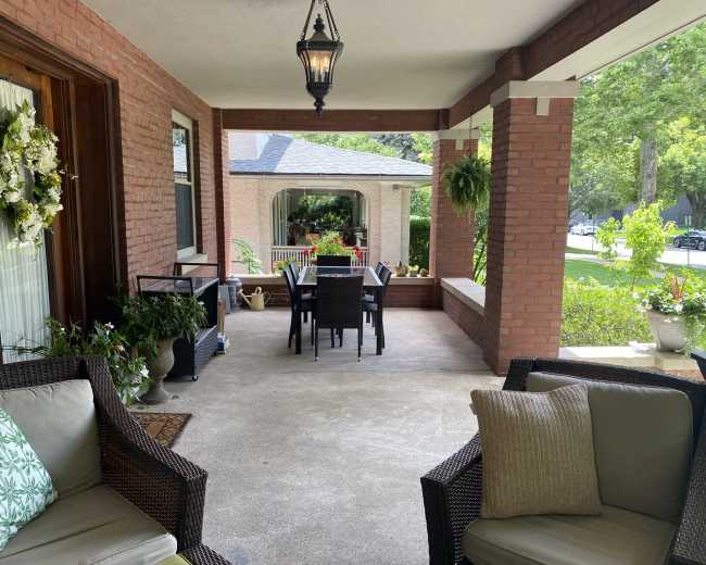 The image shows a covered porch featuring a seating area with wicker chairs and a dining table set up for four, surrounded by greenery and decorative plants.