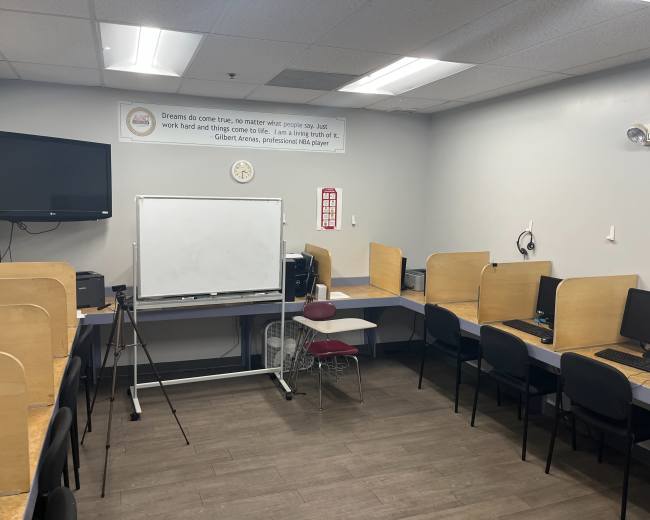 The image shows a small classroom or workroom with several computer stations, a whiteboard, and a television mounted on the wall.