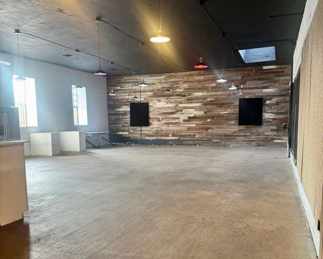 The image shows an empty interior space with a wooden accent wall, overhead pendant lighting, and a concrete floor.
