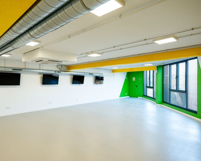 The image shows a spacious, well-lit room with green and white walls, several mounted screens, and large windows.