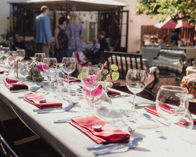 A long dining table is set with red napkins, glassware, and floral centerpieces in an outdoor setting.