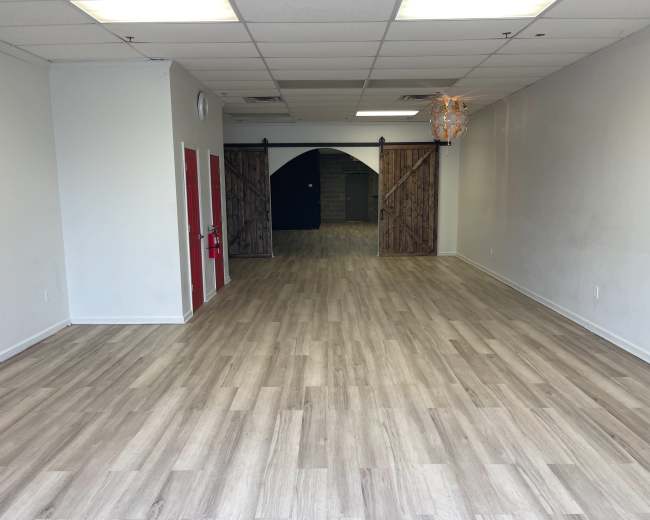 The image shows an empty retail space with wooden flooring, two large wooden doors at the back, and neutral-colored walls.