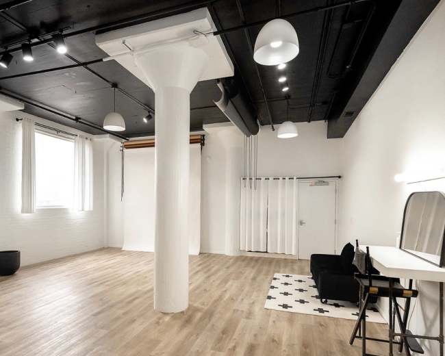 The image shows a spacious, minimalist room featuring a central white column, wooden flooring, a large mirror, and simple furniture against a white backdrop.