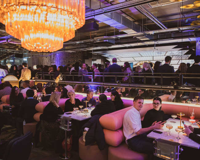 A crowded restaurant features people seated at tables and congregated in groups, with dim lighting and a stylish decor.