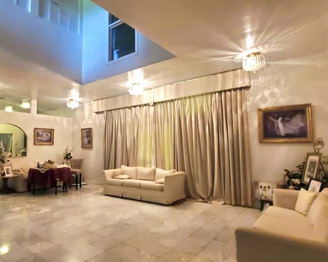 The image shows a spacious living room with a high ceiling, marble flooring, soft seating arrangements, and large curtains beside windows.