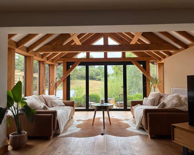 The living room features two brown sofas facing a coffee table, with large windows framed by wooden beams that provide a view of the outdoor landscape.