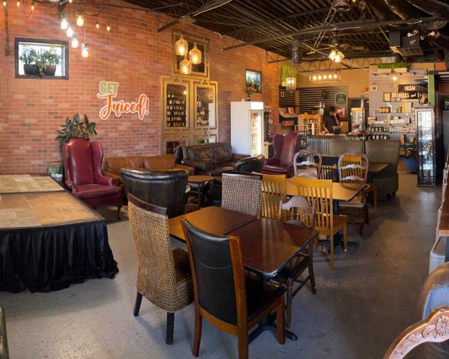 The image shows a modern café or lounge area with various seating arrangements, including armchairs, tables, and a small stage, against a brick wall backdrop.
