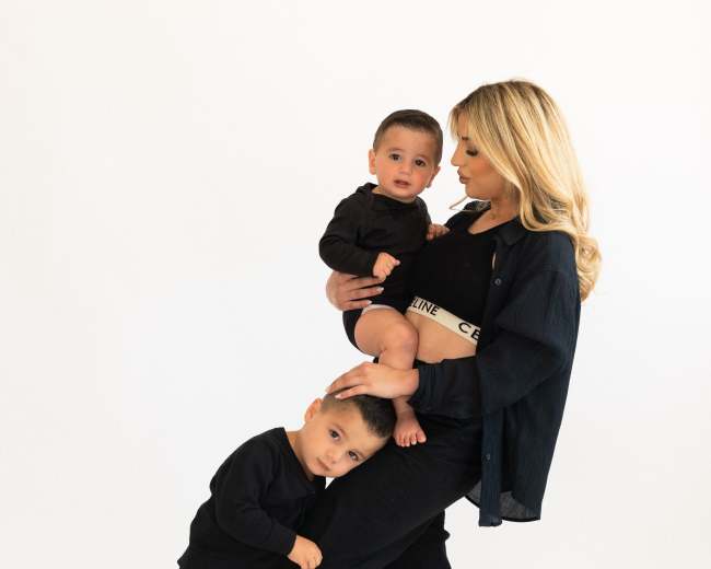 A woman stands with two children, one on her hip and the other holding her leg, all dressed in black against a white background.