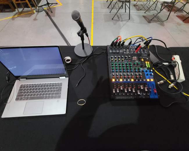 A laptop, a microphone, and an audio mixer are set up on a black table in a room with chairs arranged in the background.