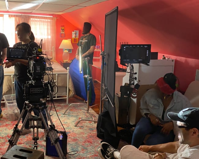 A film crew sets up lighting and equipment in a room with a bed and colorful decor for a shoot.