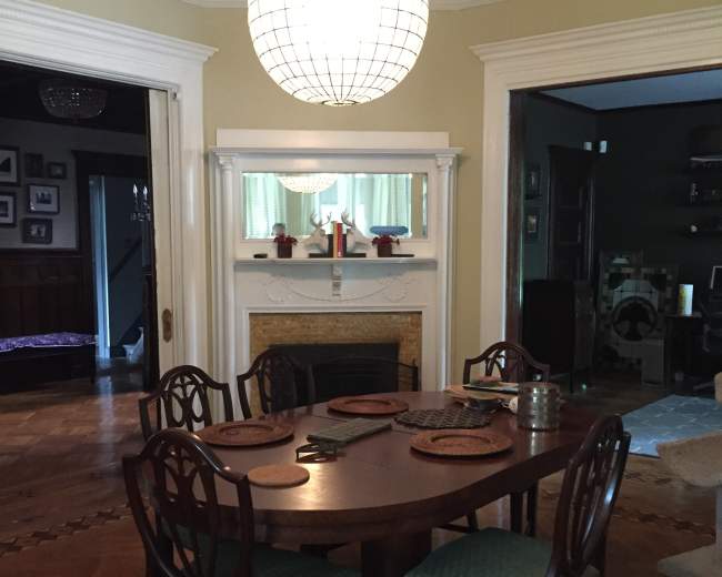 A round wooden dining table is set in a well-lit room, surrounded by four chairs, beneath a large pendant light.