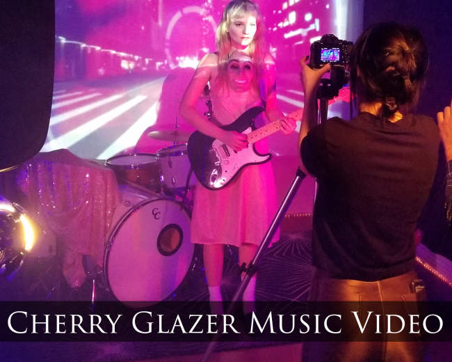 A musician performs on stage in front of a projected cityscape while being filmed by a camera operator.