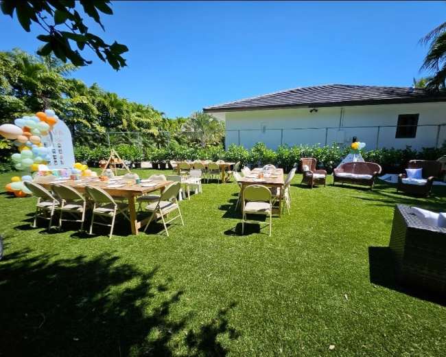 The scene shows an outdoor setup with tables and chairs arranged on green grass, surrounded by trees and a house in the background.