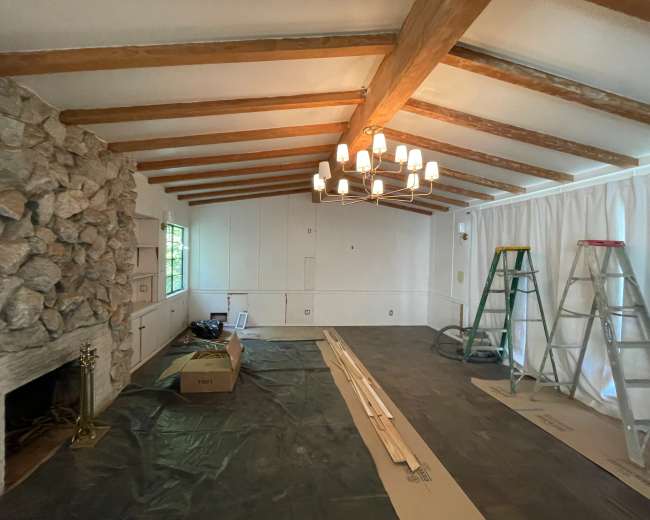 The room features exposed wooden beams on the ceiling, a stone fireplace, and bare walls with construction materials scattered on the floor.