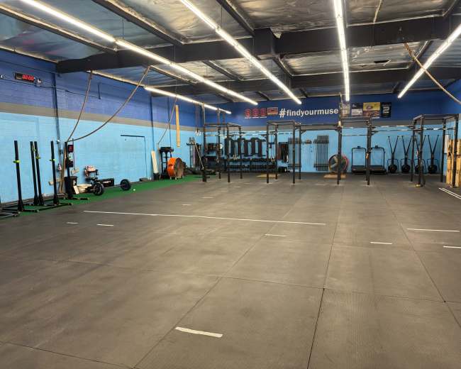 The image shows a fitness gym with various workout equipment, including weightlifting racks and exercise machines, set on a rubber flooring surface.
