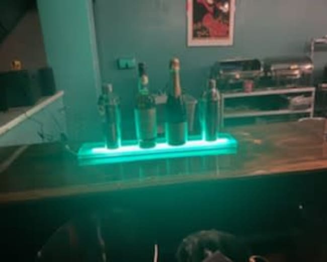 The image shows a bar countertop with four bottles and a cocktail shaker illuminated by a glowing turquoise light.