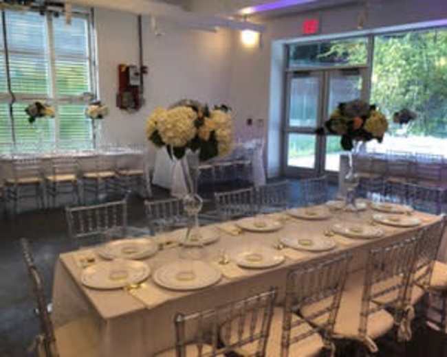 A banquet hall features elegantly set tables with white tablecloths, ornate place settings, and floral centerpieces, illuminated by natural light from large windows.