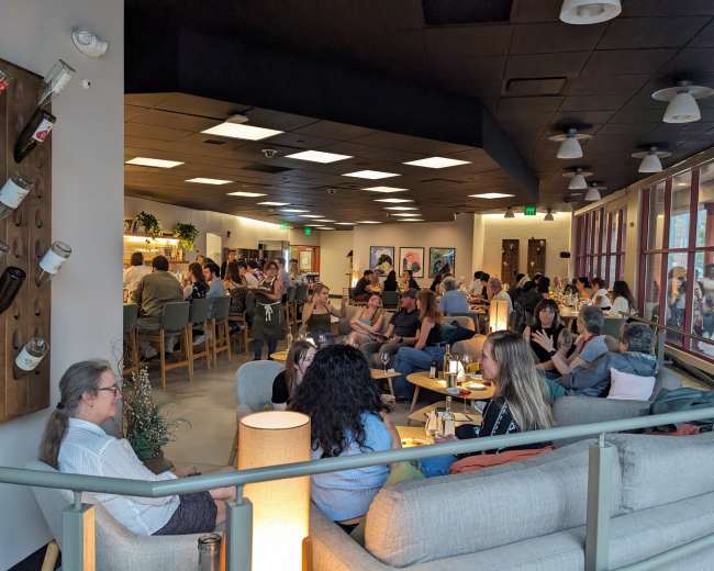 The image shows a crowded cafe interior with patrons seated at various tables engaged in conversation, while others are at the bar.