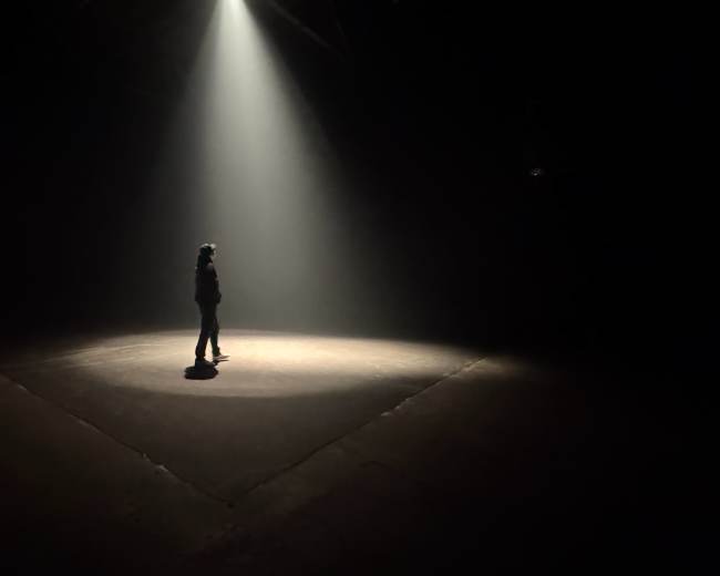A solitary figure stands in a beam of light on a dark stage.