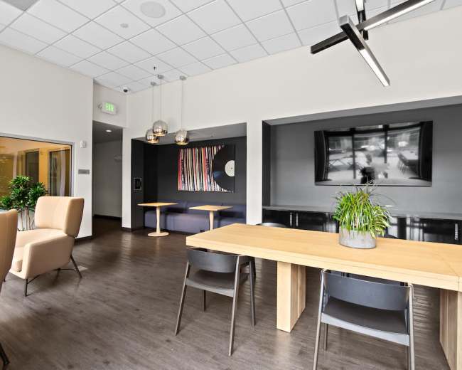 The image shows a modern interior space with wooden tables, stylish chairs, and decorative lighting, featuring a wall-mounted television and abstract artwork.