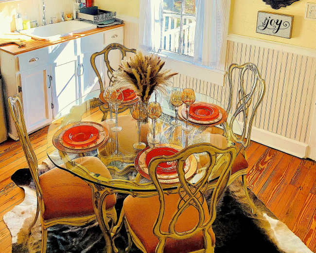 A dining area features a glass table with orange plates and decorative glassware, surrounded by elegant chairs, with a window and kitchen cabinetry in the background.
