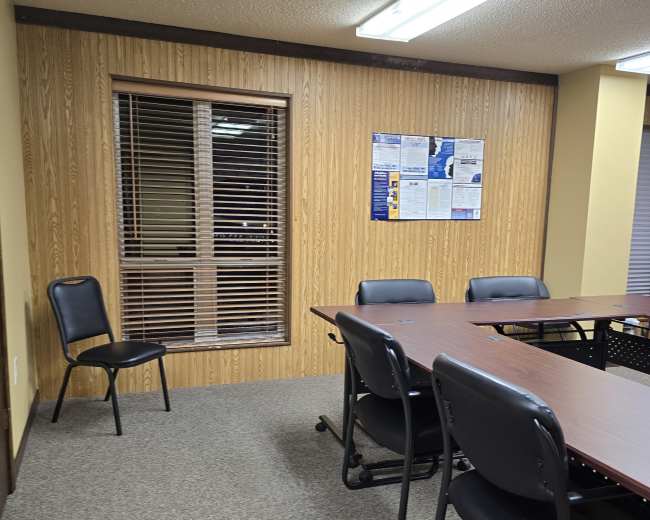 A conference room with a large table surrounded by chairs and a single empty chair against a wooden-paneled wall with a window.