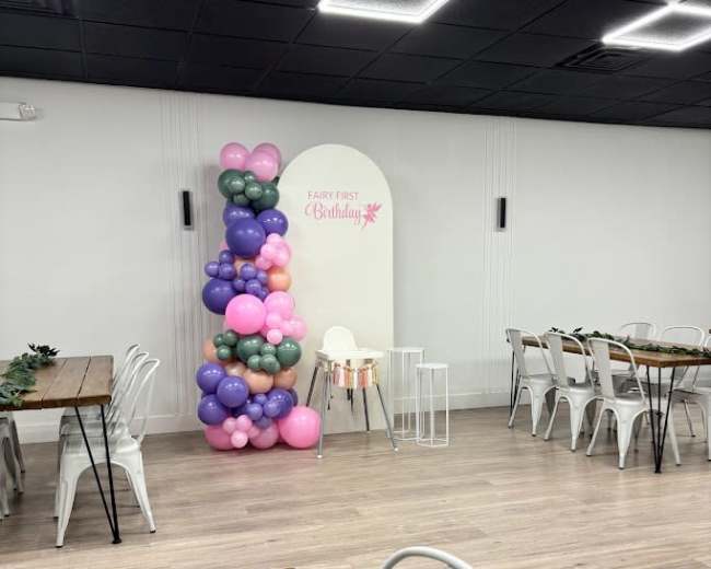 The image shows a decorated indoor party space featuring a balloon arrangement in various colors, a table setting, and chairs arranged for a birthday celebration.