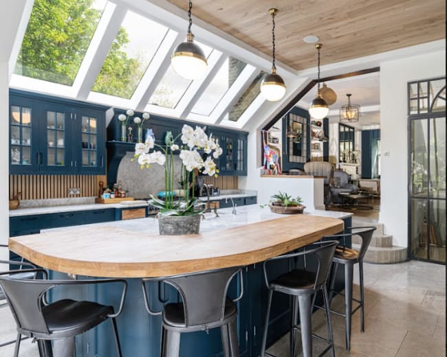 The image shows a modern kitchen with a large wooden island, blue cabinetry, and skylights allowing natural light to fill the space.