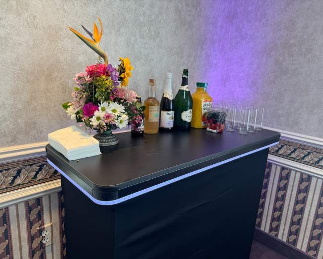 A bar setup is displayed against a textured wall, featuring various bottles of drinks, a floral arrangement, and several empty glasses.